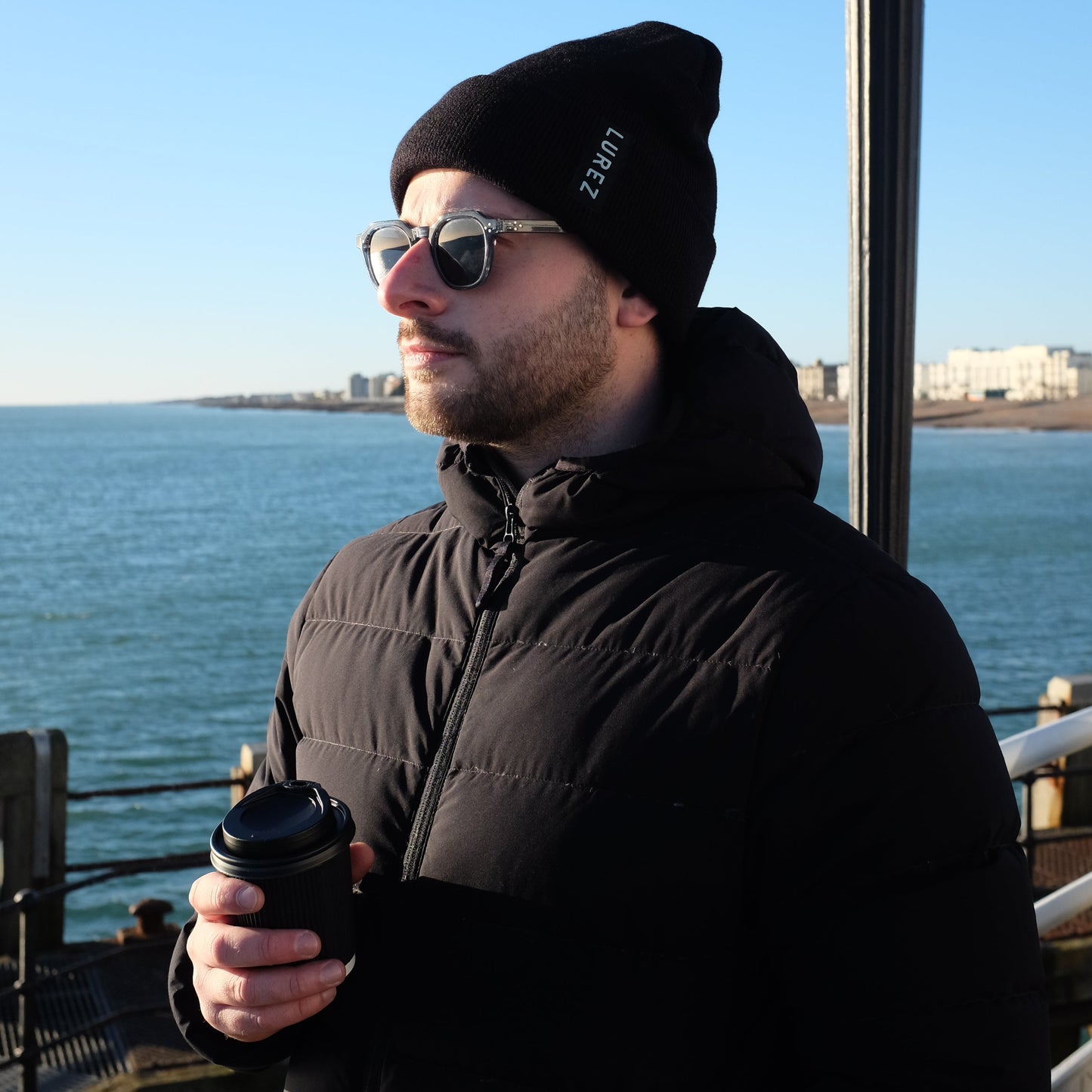 Man wearing a black beanie and sunglasses by a waterfront with a clear blue sky.