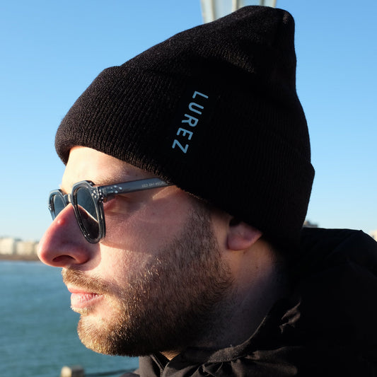 Man wearing a black beanie with 'LUREZ' branding, sunglasses, and a black jacket by a body of water.